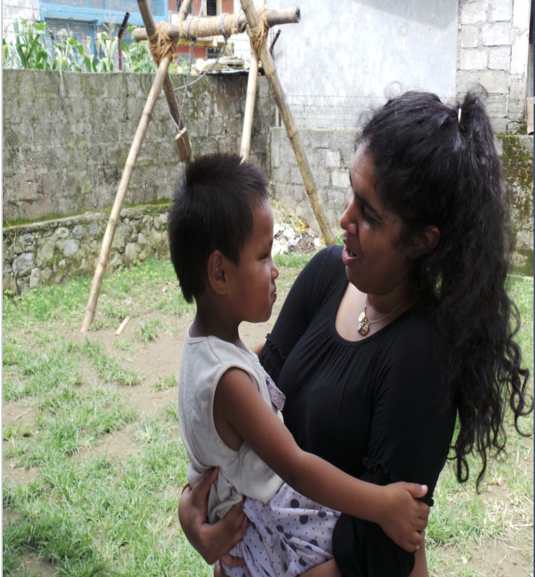 A woman holding a young girl in her arms outside in a grassy area, with a wooden swing in the background and a stone wall and crops behind them.