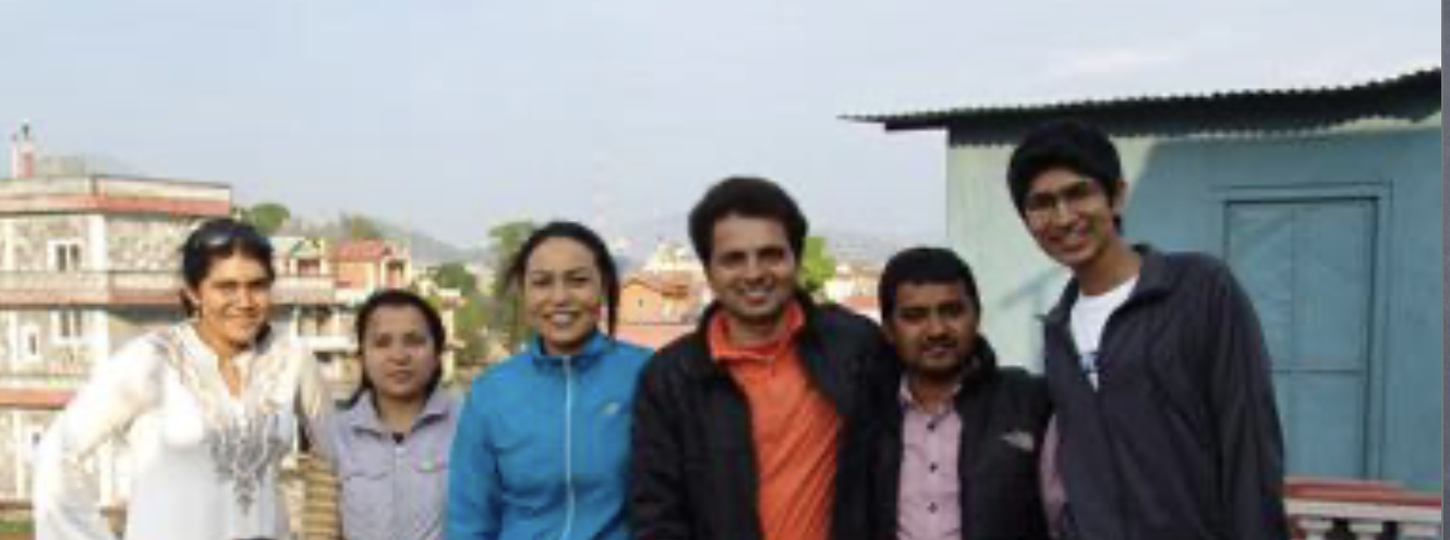 Group of six people standing outdoors in front of buildings and a small structure, smiling at the camera.