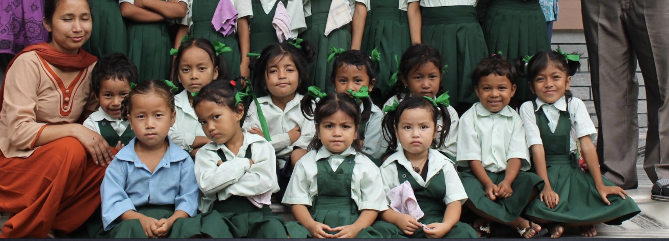 A group of young girls and a woman, likely a teacher, posing for a class photo. The girls are wearing school uniforms with green skirts and light-colored shirts, some with green ribbons in their hair. The woman is dressed in traditional clothing with
