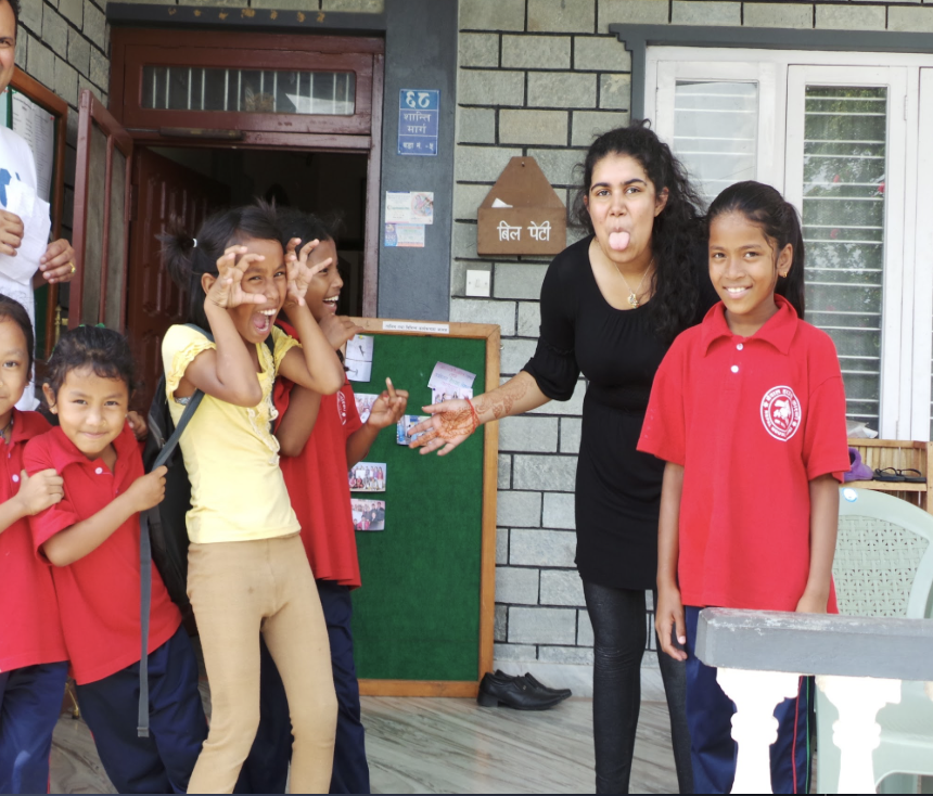 A group of children and a woman standing outside a building, smiling and making playful faces. The woman is sticking out her tongue and gesturing as the children appear amused.