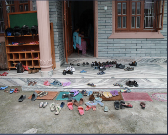 Multiple pairs of shoes and slippers are arranged outside the entrance of a house, with a child sitting at the door.