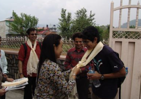 A woman in a floral blouse is tying a white scarf around the neck of a young man in a black shirt, while three other men look on. They are outdoors near a white fence, with trees and houses in the background.