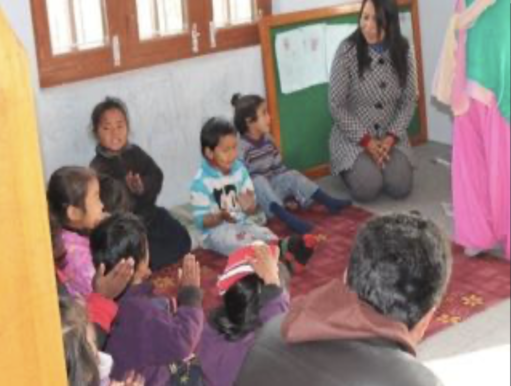 Children sitting on the floor with hands folded in prayer, a woman kneeling in front of them, in a classroom setting.
