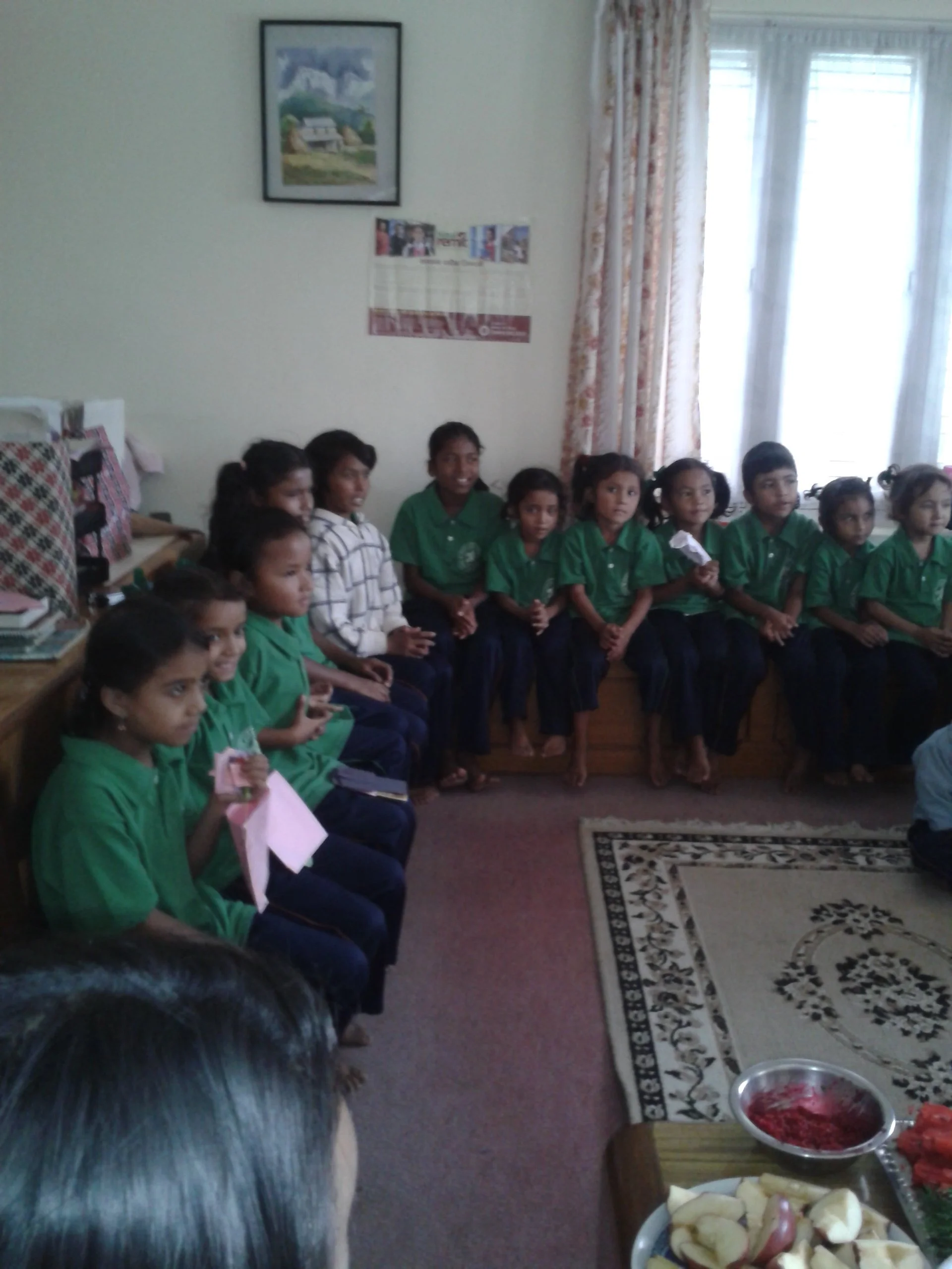Group of children sitting on a bench inside a room, some holding papers, with a table of food in the foreground.