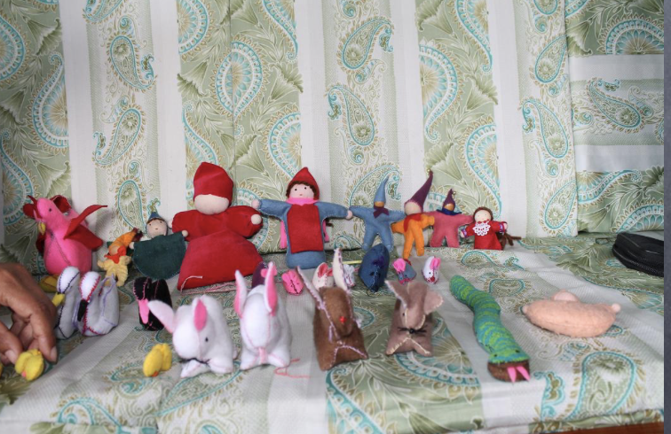 Collection of handmade stuffed dolls and animals arranged on a table with a paisley-patterned curtain background.