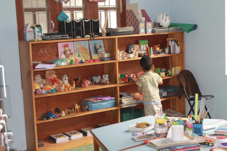 A young boy in a yellow plaid shirt searches through a large wooden bookshelf filled with toys, books, and art supplies in a classroom.