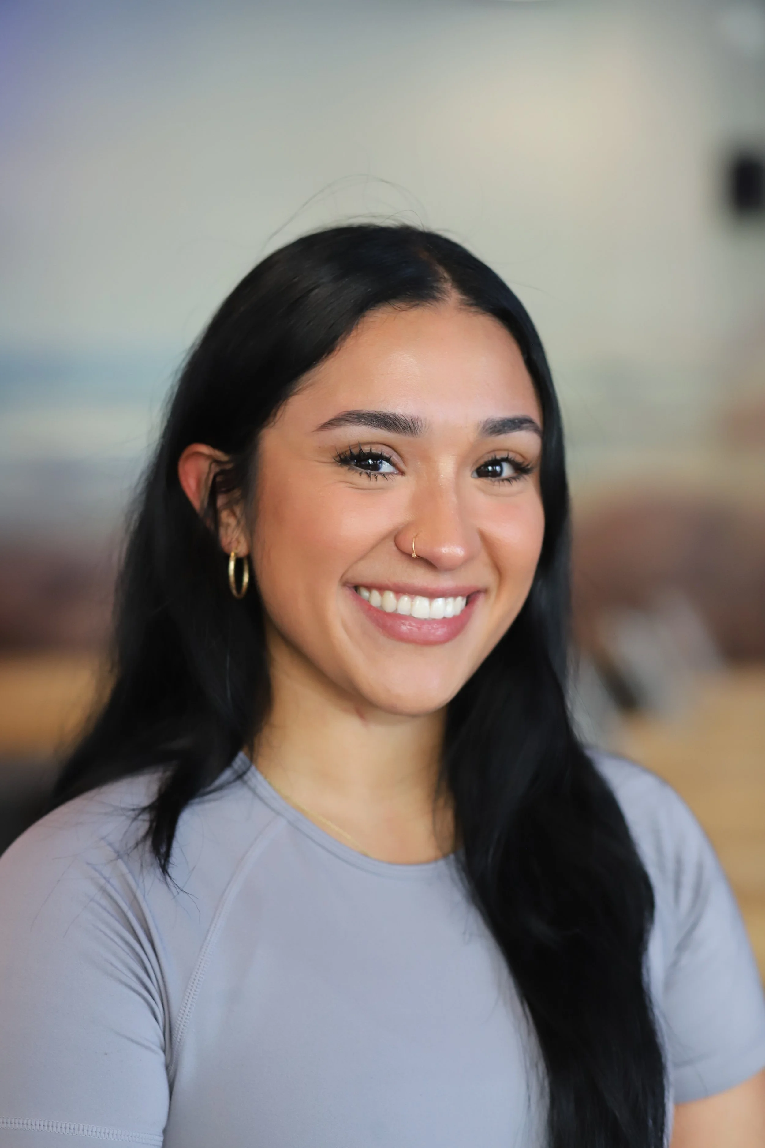 Smiling young woman with long black hair, hoop earrings, and a nose ring, wearing a light gray shirt, indoors with blurred background.