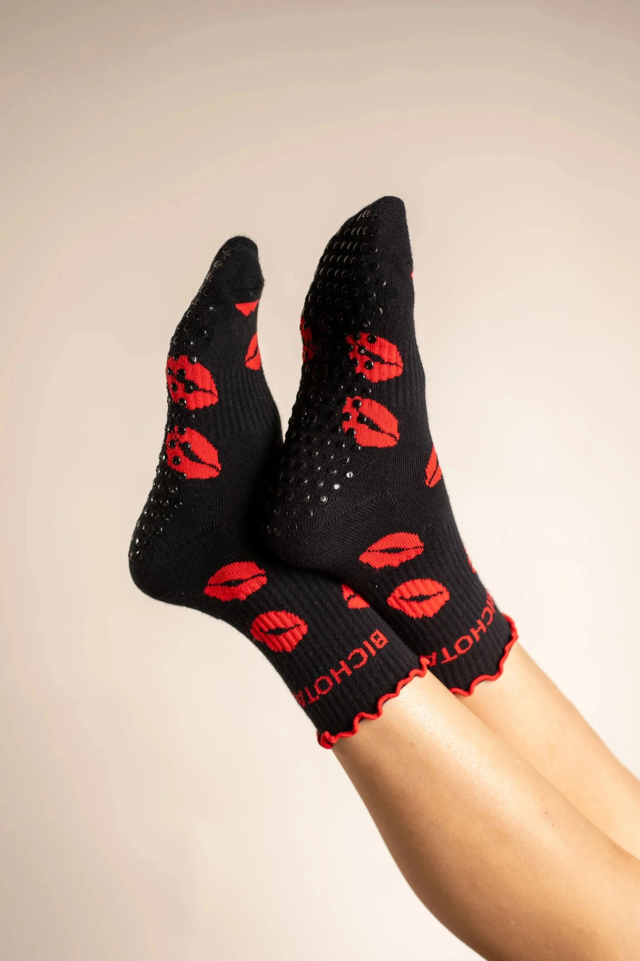 A pair of black socks with red lip print designs and small black rhinestones, worn on feet with legs up against a plain background.