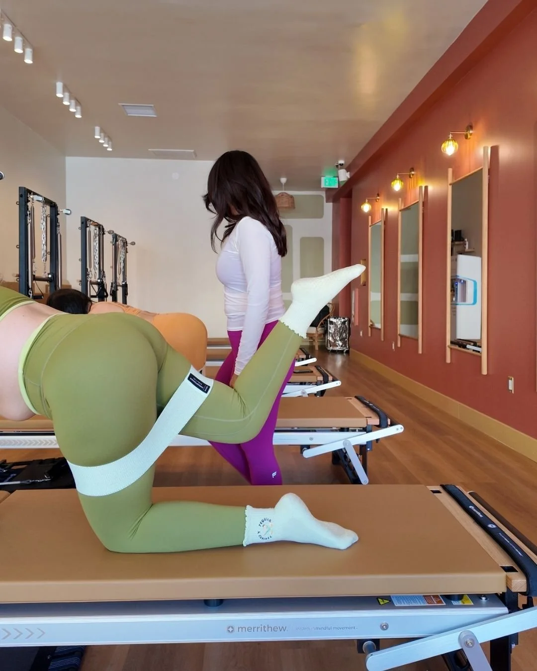 A woman doing a Pilates exercise on a reformer machine in a fitness studio with mirrors and plants.