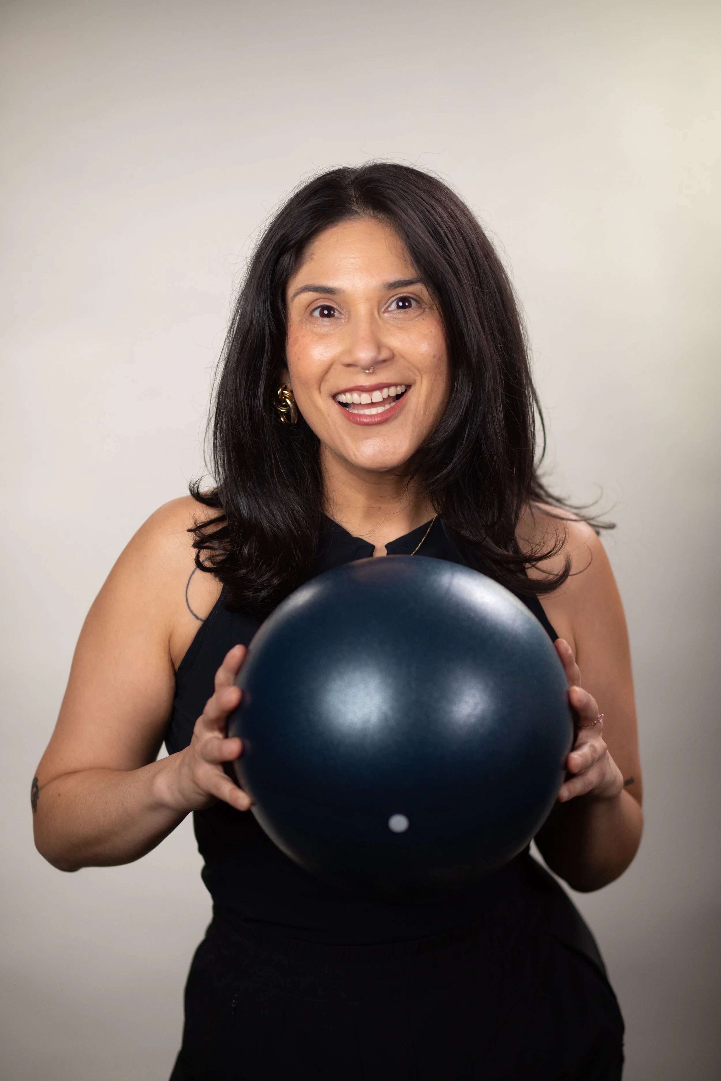 Smiling woman holding a black exercise ball, wearing a black sleeveless top and gold earrings, against a neutral background.