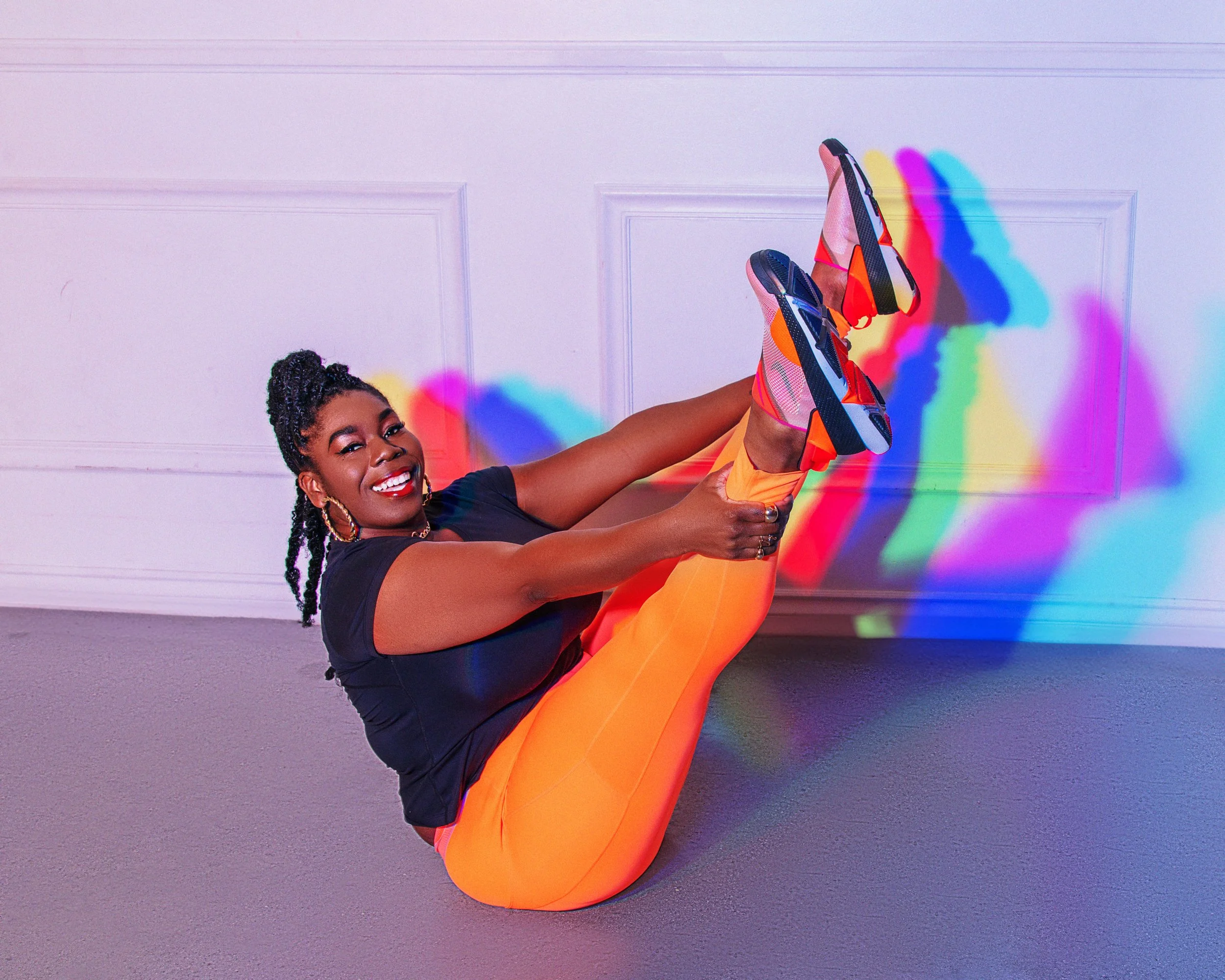 A woman with braided hair and hoop earrings in workout clothes performing a stretch or yoga pose on the floor, with colorful rainbow light shadows cast on the wall behind her.