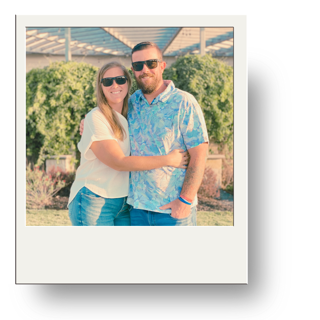 A smiling couple wearing sunglasses standing outdoors, embracing each other with green bushes and a structure with glass panels in the background.