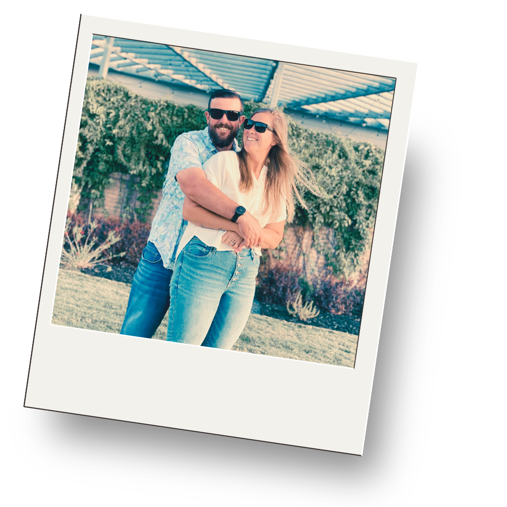 A smiling man with a beard and sunglasses hugging a smiling woman with sunglasses outdoors in a garden with plants and a structure overhead.