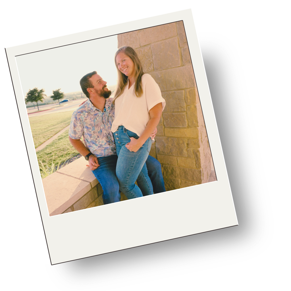 A man with a beard and a floral shirt sitting on a concrete bench, looking up and smiling at a woman standing beside him. The woman has long hair, wearing a white shirt and blue jeans, smiling back at the man. They are outside near a brick wall and trees under a bright sky.