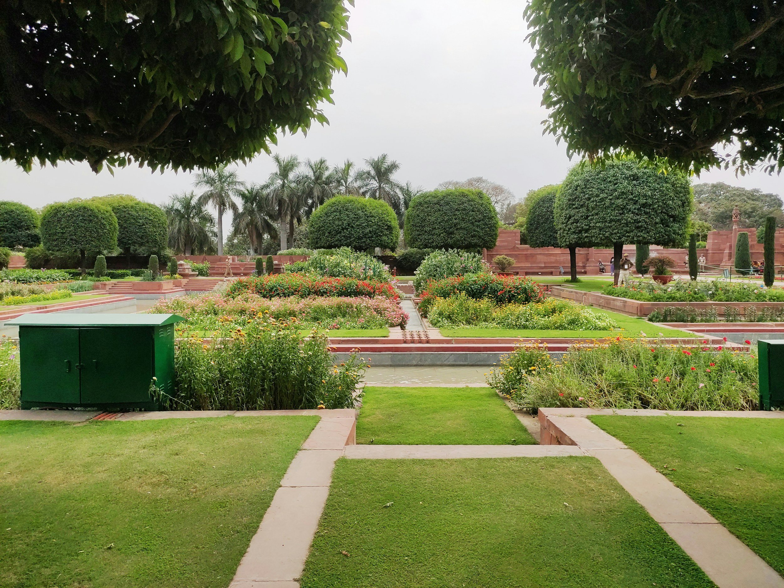 A well-maintained garden with neatly trimmed trees, vibrant flowers, and paved walkways. Large trees with rounded foliage and a variety of plants are visible under an overcast sky.