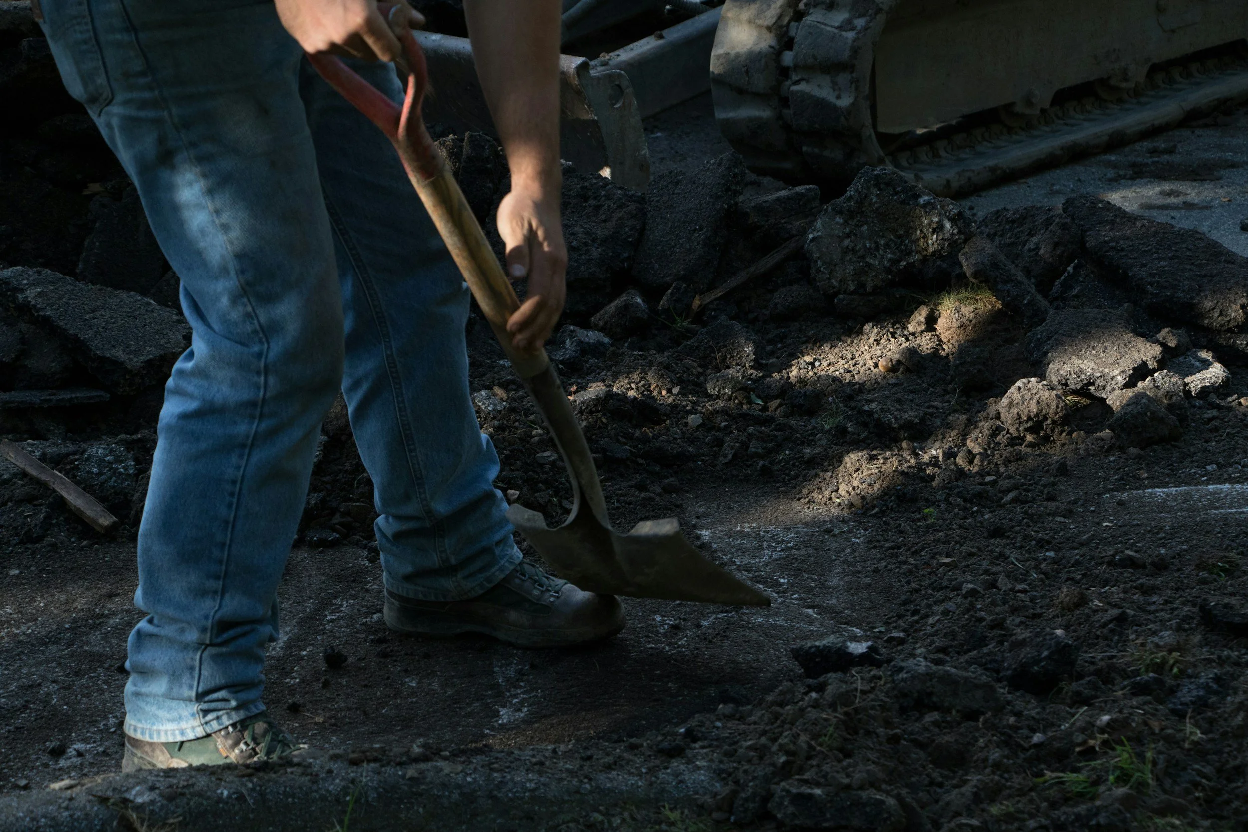 Person digging in dark soil with a shovel, with construction equipment in the background.