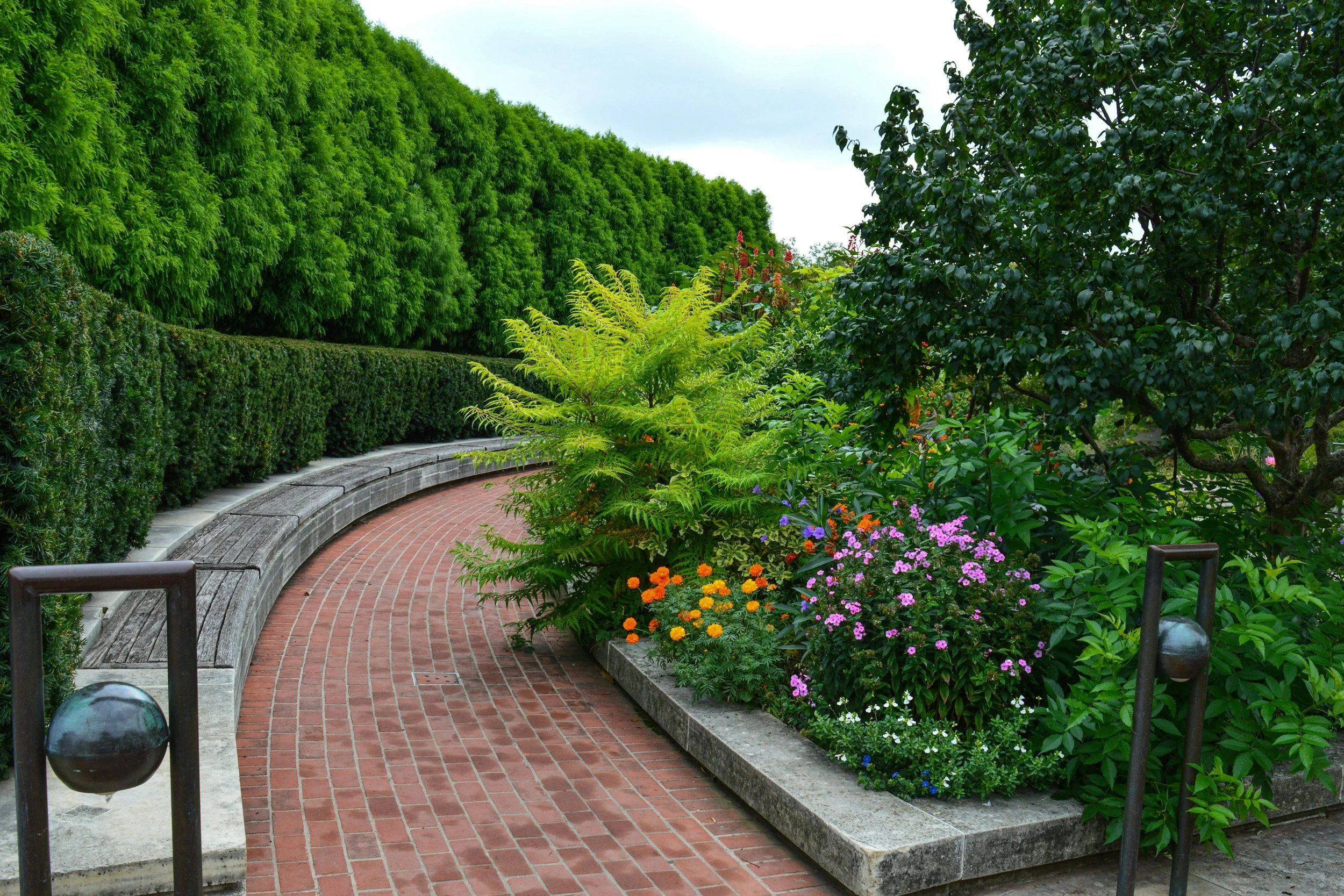 Curving brick pathway through a lush garden with various green bushes and colorful flowers, bordered by a low stone edge and black metal posts.