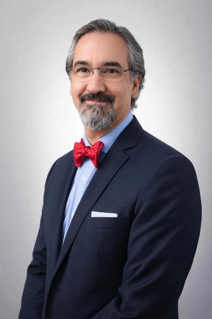 Hombre con barba y gafas, vestido con traje azul oscuro, camisa azul y pajarita roja con puntos blancos, posando frente a fondo gris.