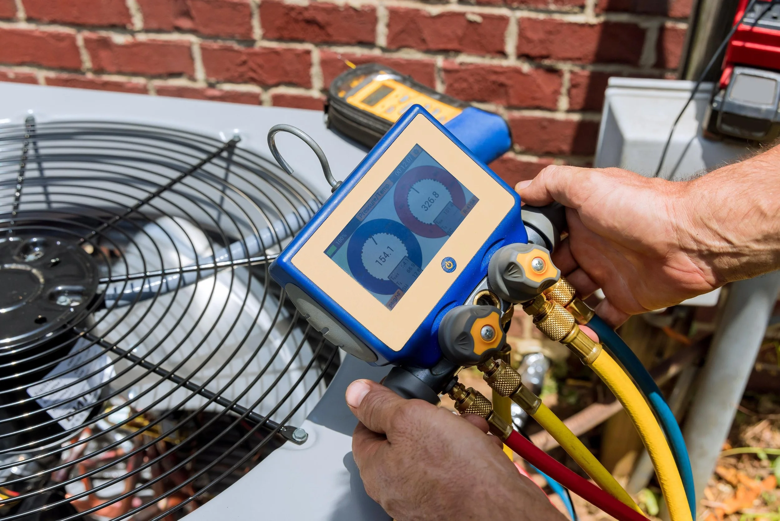 Technician using a digital manifold gauge on an outdoor air conditioning unit, with a wire fan and brick wall in the background. HVAC repair and diagnostics. Lafollette Jacksboro Caryville East Tennessee Refrigerant 