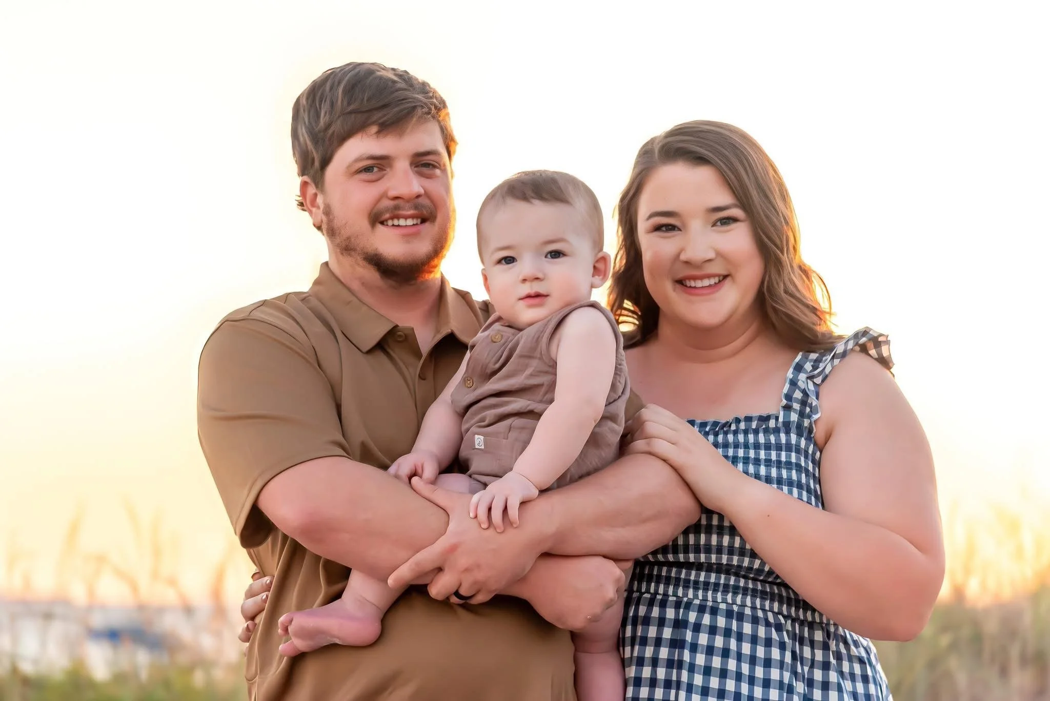 A family of three smiling outdoors at sunset, with the father holding a small child and the mother standing beside them. Local East Tennessee HVAC repair and maintenance
