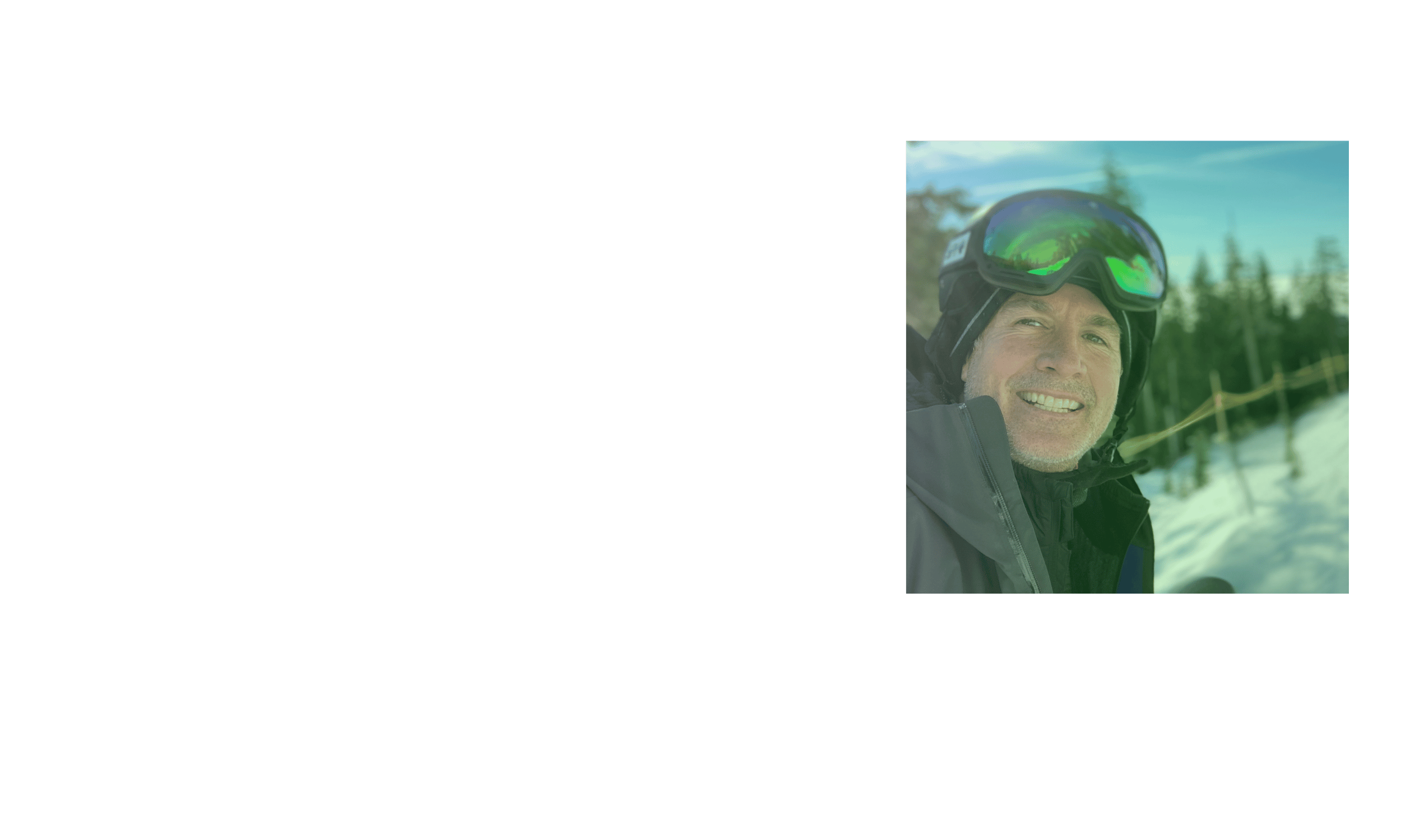 A man wearing snow goggles and a winter jacket smiling outdoors on a snowy day with trees and a ski slope in the background.