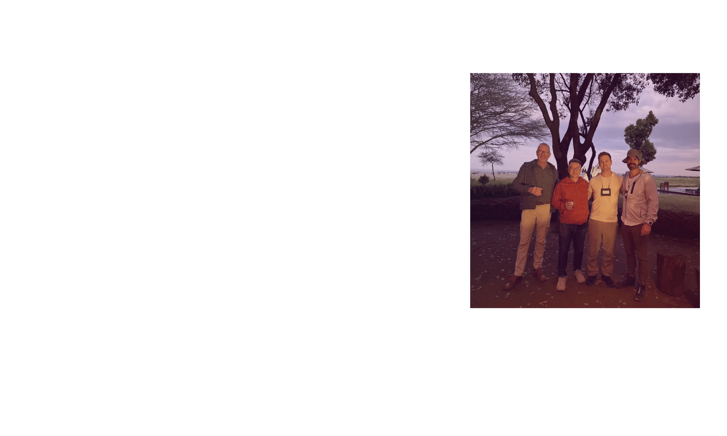 Group of four men standing outdoors at sunset, with a large tree and open landscape in the background, some holding drinks and smiling.