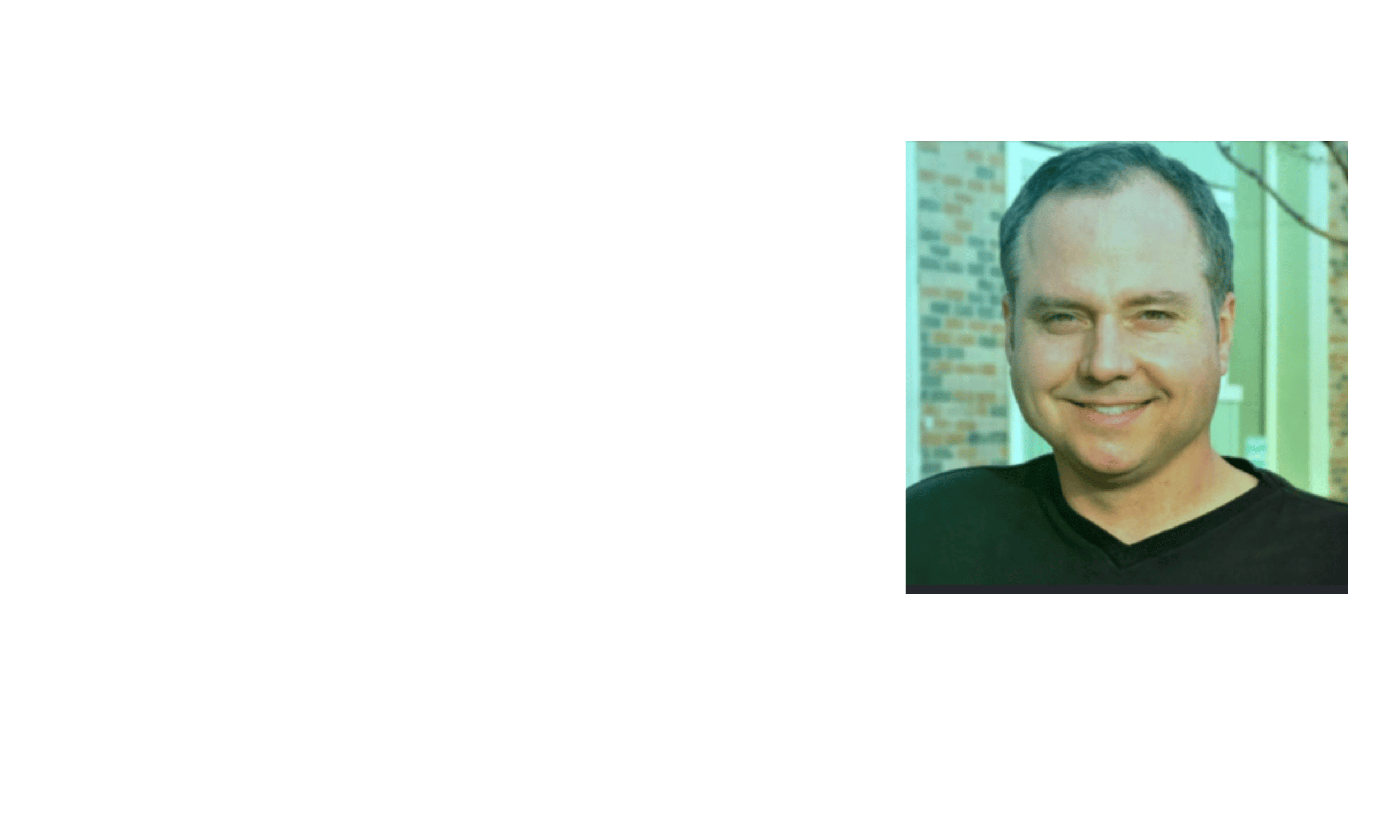 A smiling man in a black shirt standing outdoors in front of a building with brick walls and glass windows.
