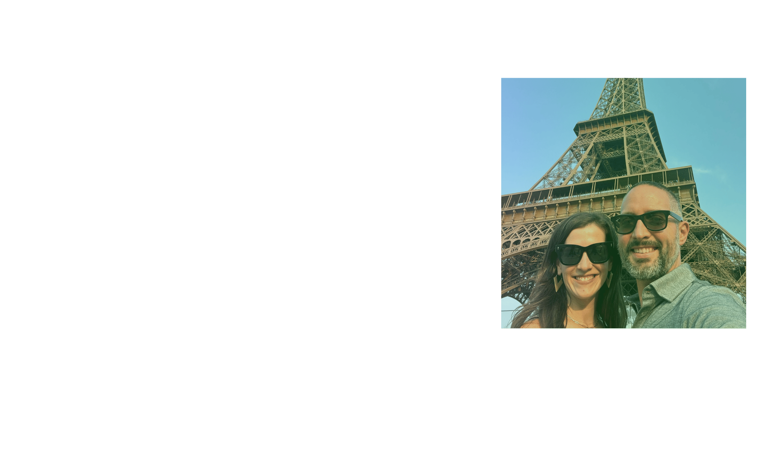 A smiling couple wearing sunglasses taking a selfie in front of the Eiffel Tower under a clear blue sky.