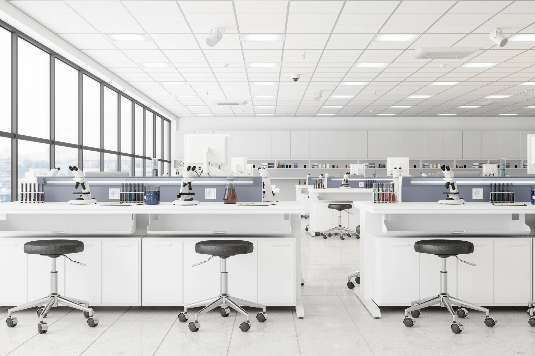 Modern science laboratory with white counters, microscopes, test tubes, and laboratory equipment. Large windows on the left let in natural light, and stools are positioned in front of workstations.