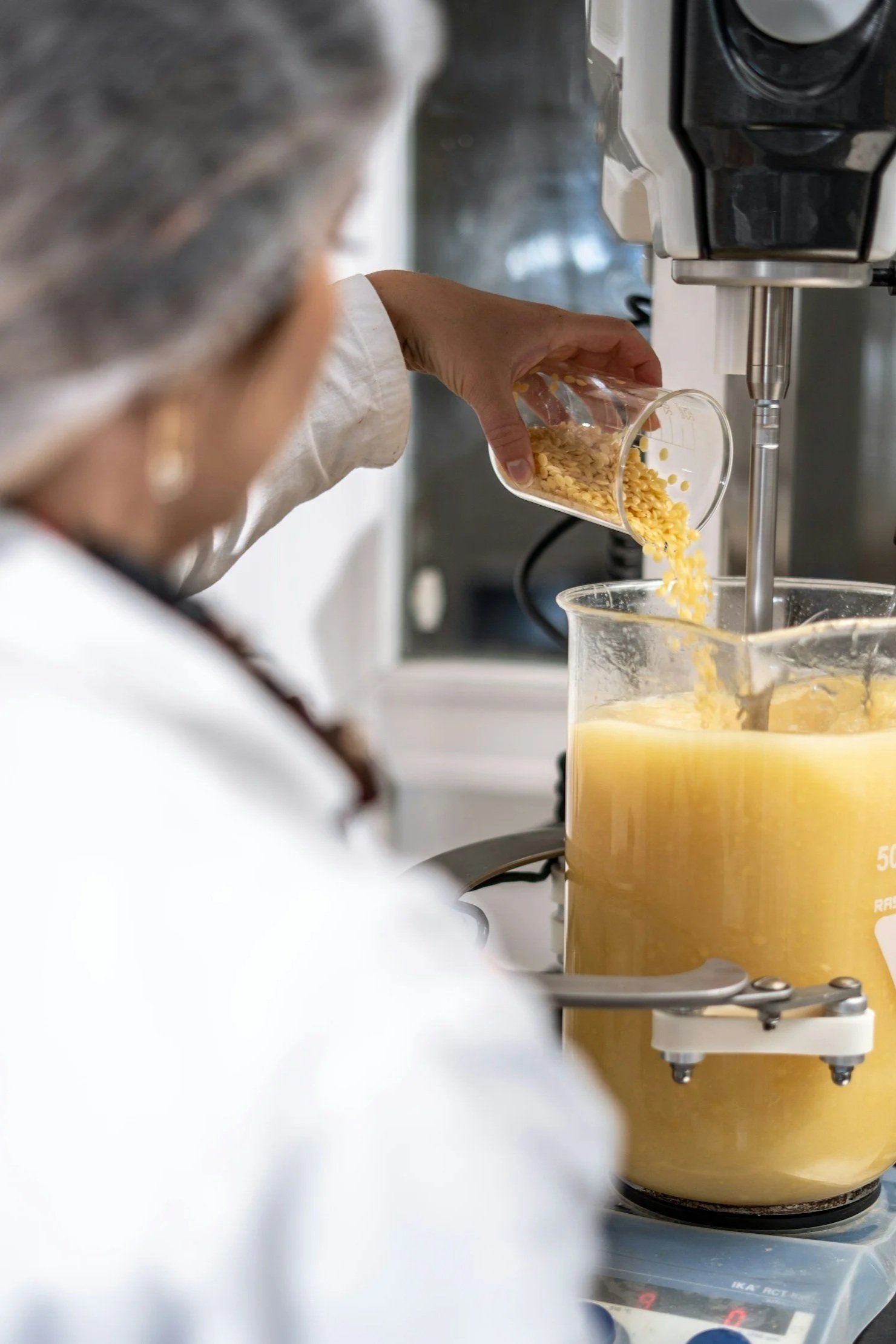 A person pouring yellow powder or grains into a large yellow liquid in a beaker on a laboratory or industrial machine.