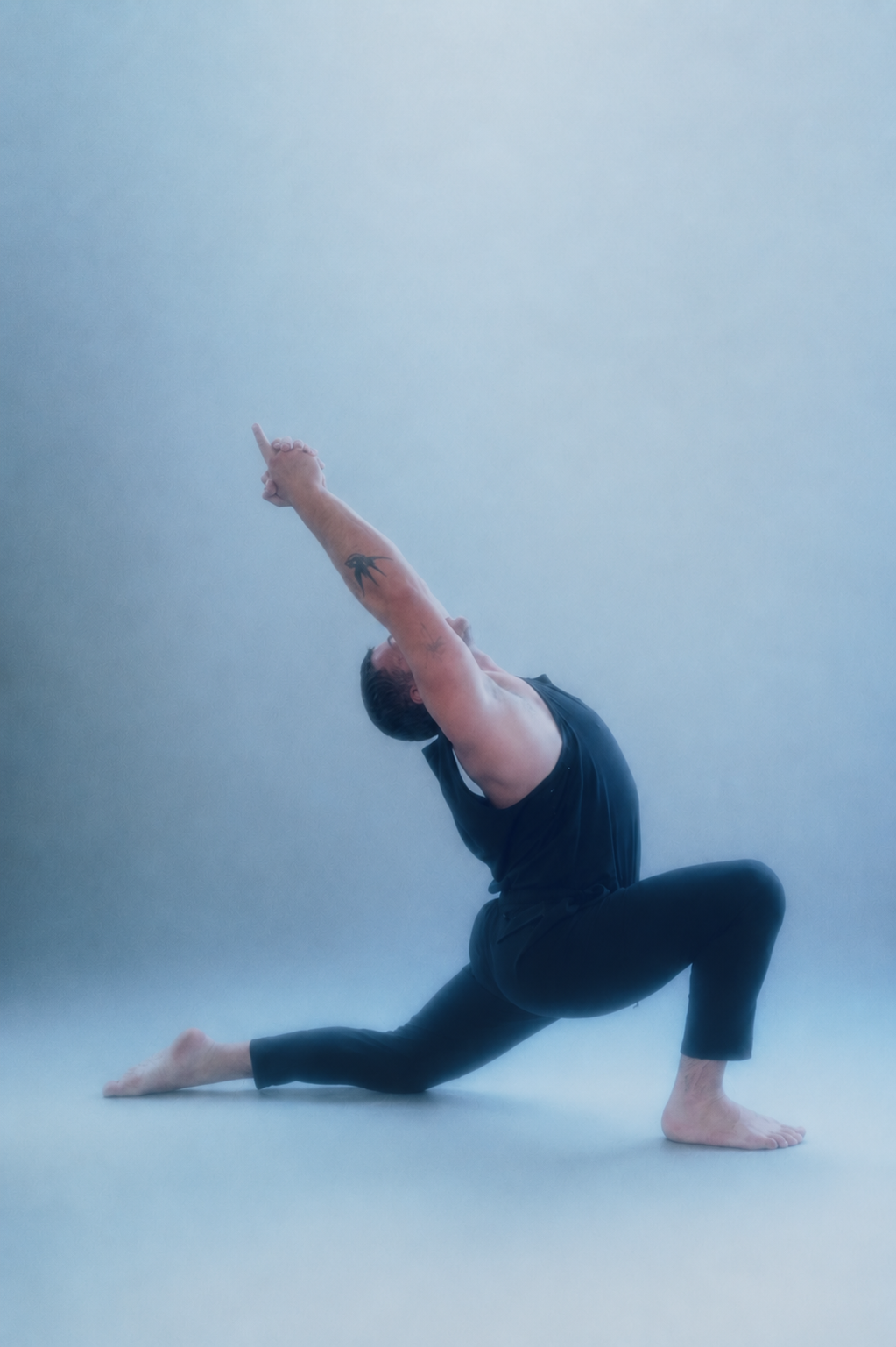 Chris Magee practicing yoga in a lunge pose with one arm extended upward and the other reaching back, wearing a sleeveless black shirt and pants, with a tattoo on his forearm, against a plain light-colored background.