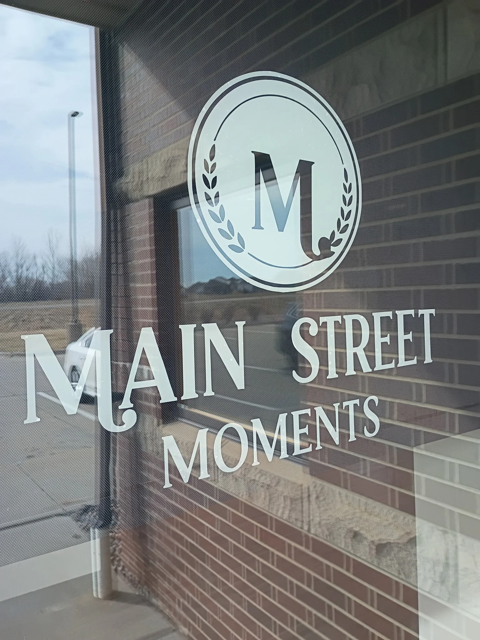 Window decal with logo and text that reads 'Main Street Moments' and a circular emblem with an 'M' and laurel branches.