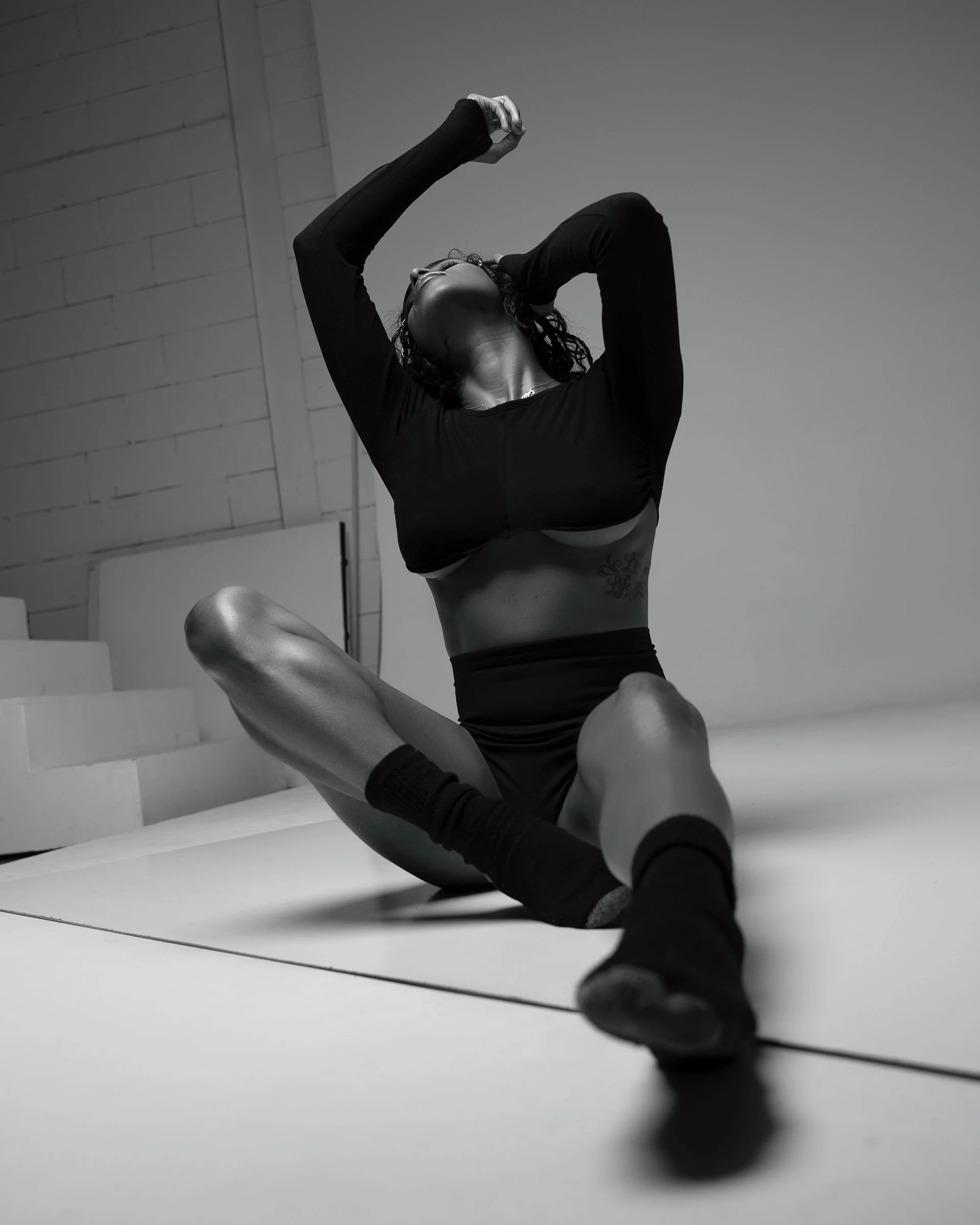 Black and white photograph of a woman sitting on a yoga mat, stretching with arms raised above her head, wearing a crop top, shorts, and socks, in an indoor studio.