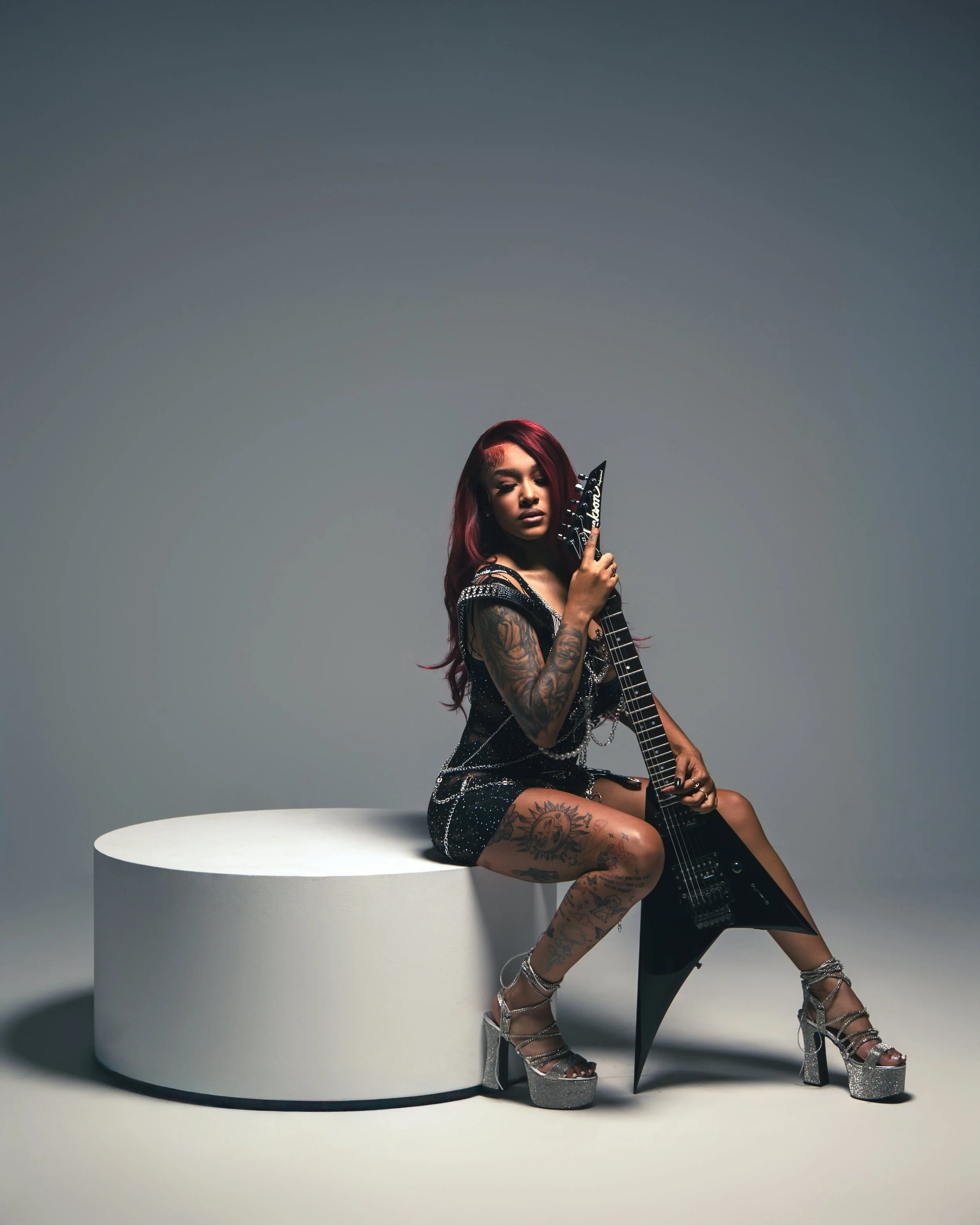 A woman with red hair and tattoos, wearing a black dress with silver details, sitting on a white circular platform, holding a black electric guitar, with a neutral gray background.