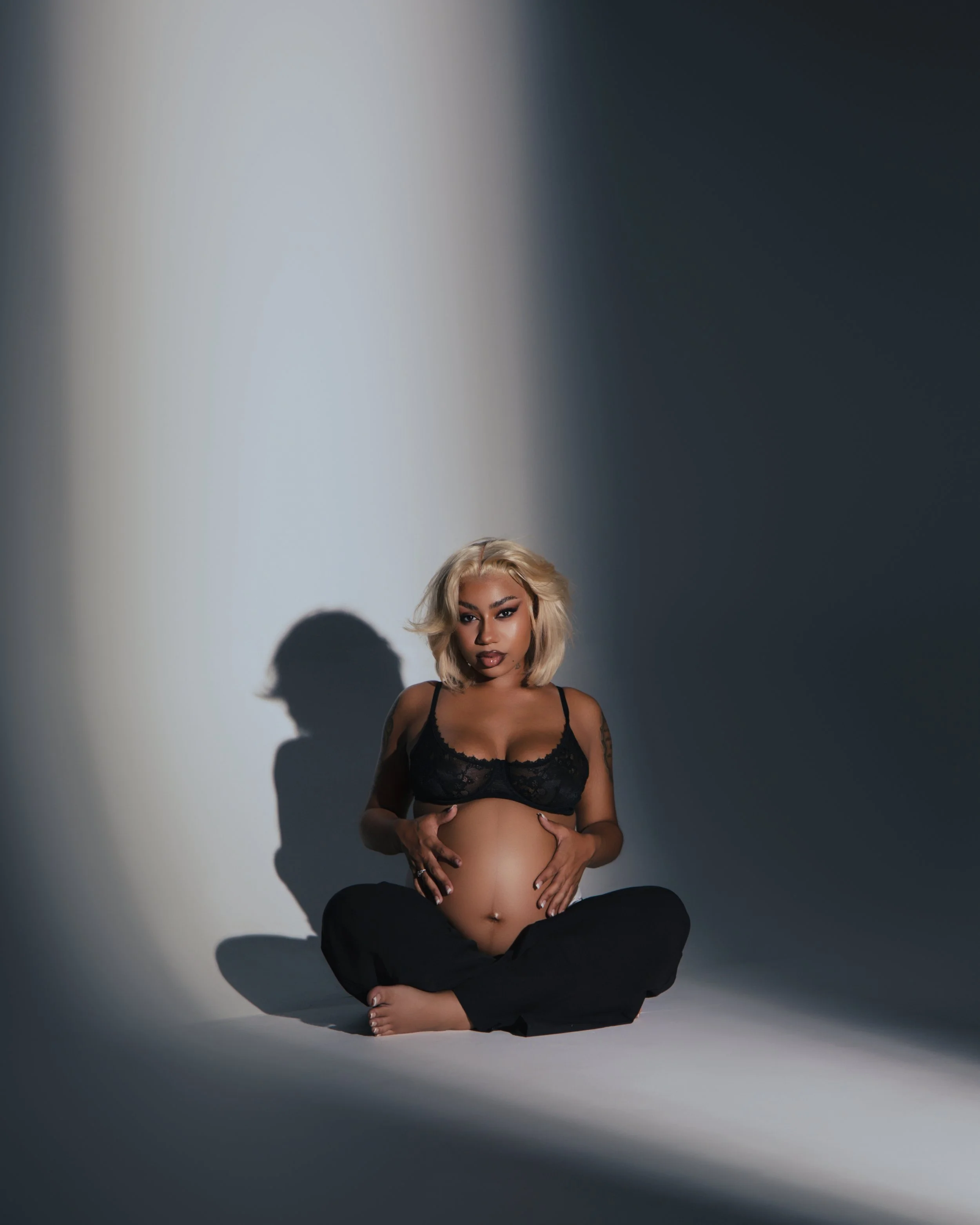 A pregnant woman with blonde hair, wearing black lace lingerie and black pants, sitting cross-legged on the floor in a studio with a gradient background. She is gently resting her hands on her belly and looking directly at the camera, with a shadow cast behind her.