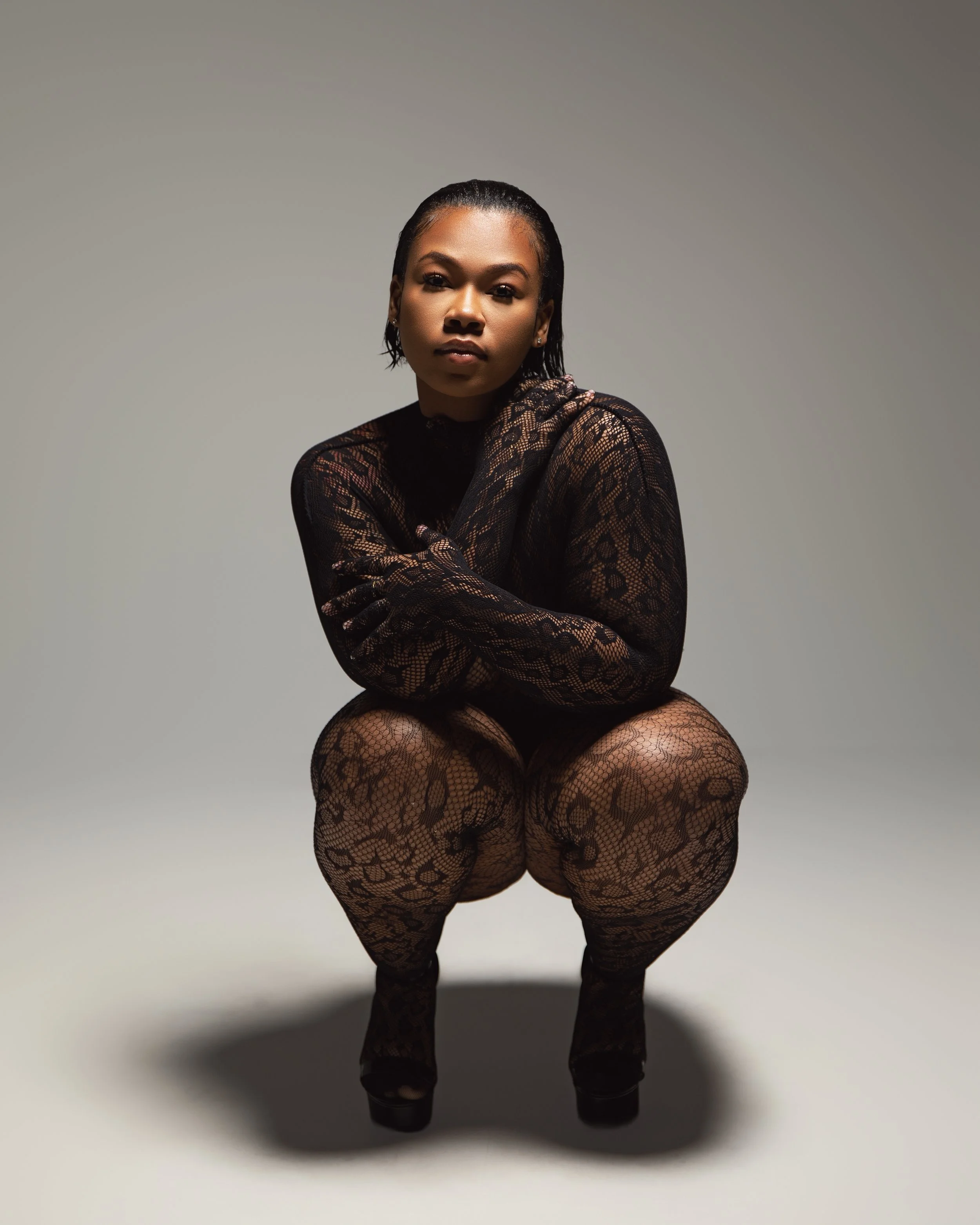 A woman in black lace clothing crouching against a gray background.