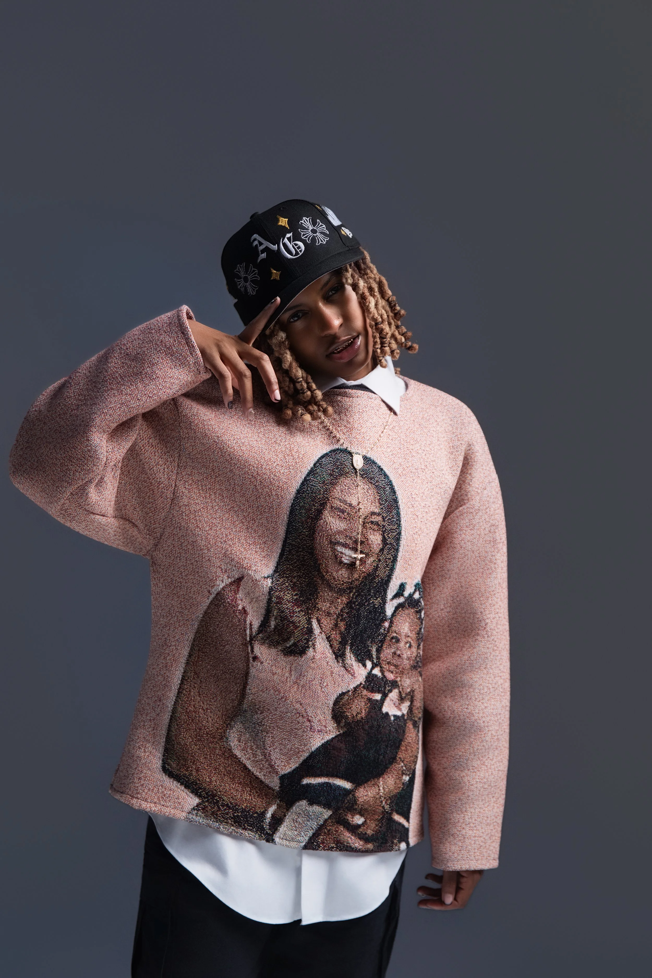 Young person with blonde twists hairstyle wearing a black cap, pink graphic sweater, and white shirt underneath, posing against a dark gray background.