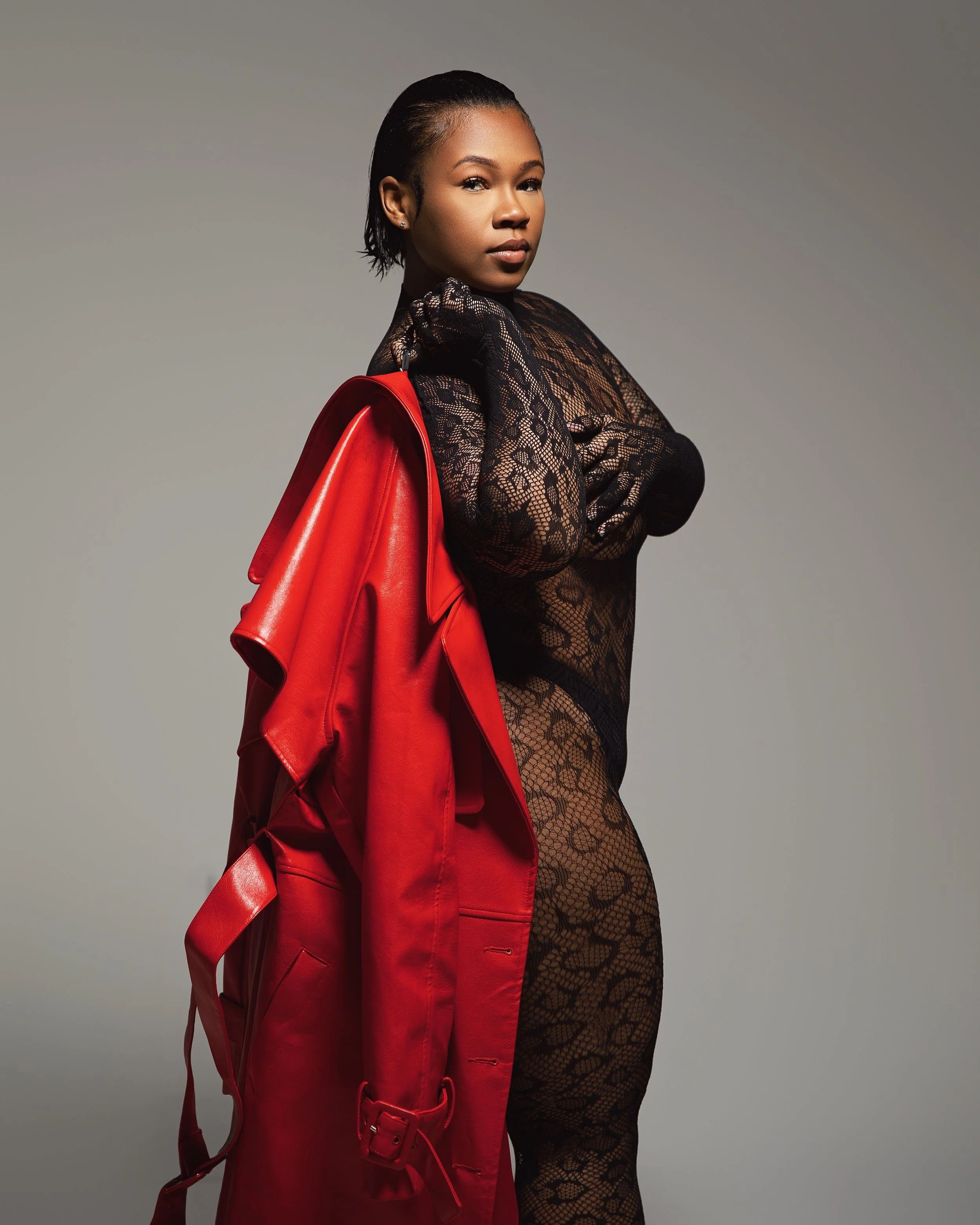 A woman in black lace clothing holding a red coat over her shoulder against a gray background.
