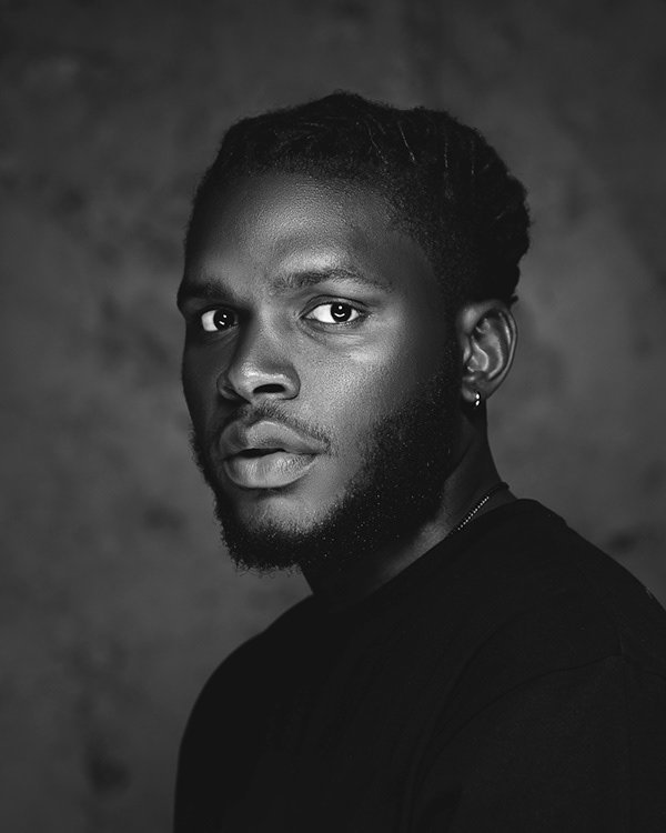 Black and white portrait of a young man with textured hair, a beard, and an earring, looking directly at the camera with a serious expression.