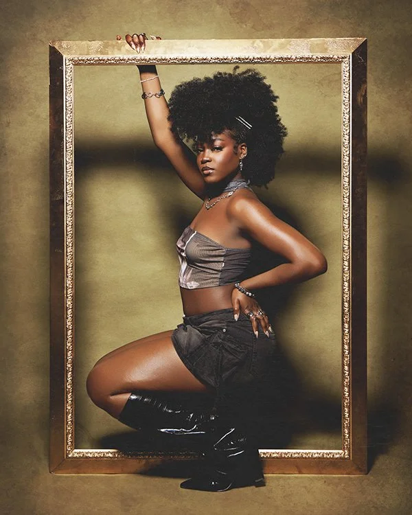 A woman with curly black hair poses inside a large wooden picture frame, wearing a metallic tube top, black shorts, and knee-high black boots, against a plain background.