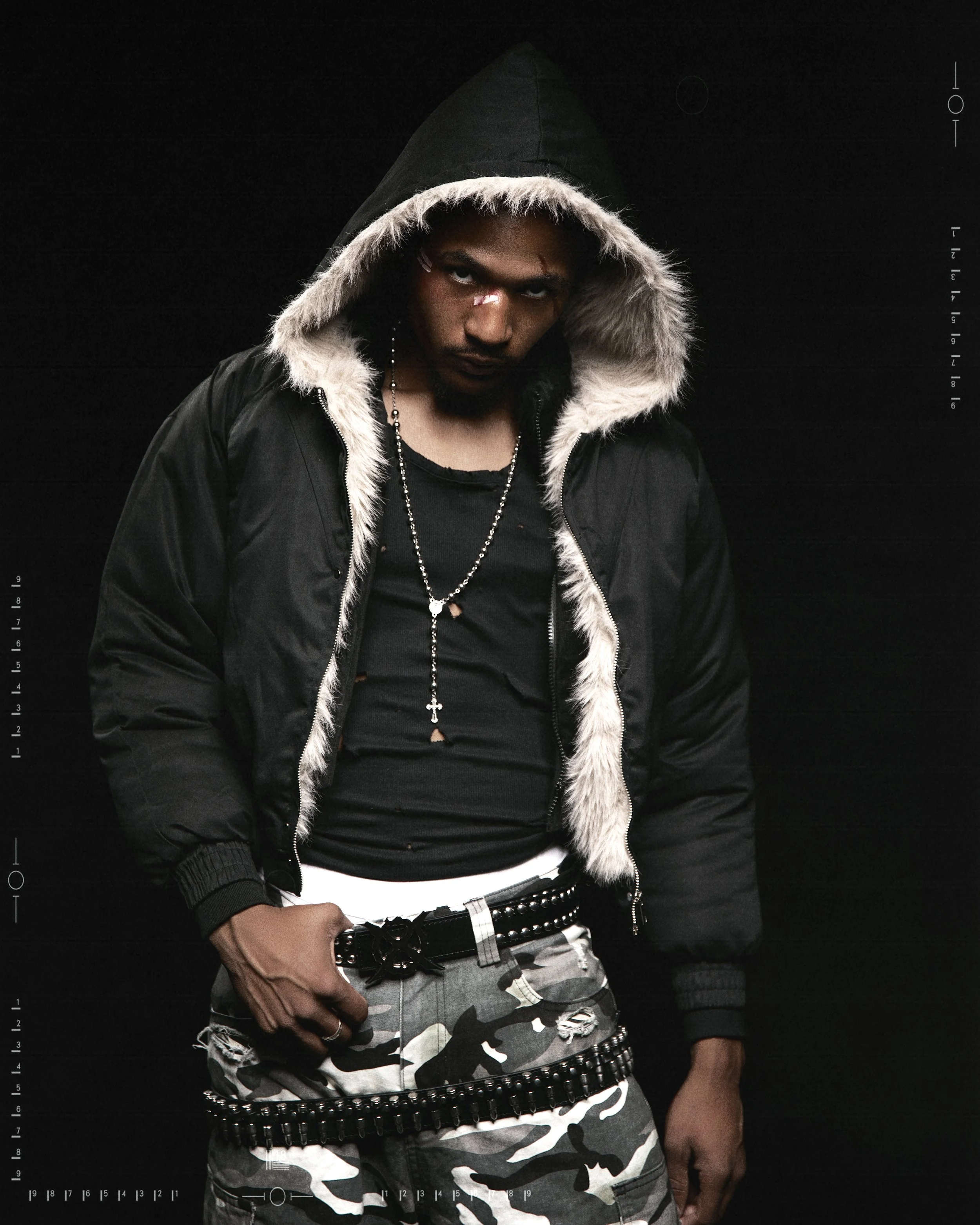 A young man with a serious expression, wearing a black hoodie with a fur-lined jacket, camouflage pants, multiple belts, and jewelry, standing against a black background.