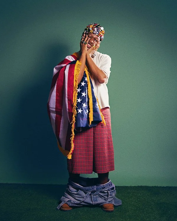 Person with colorful accessories, wearing red plaid pants and a white shirt, standing on a green surface against a plain green background, with large pants pulled down to reveal sandals.