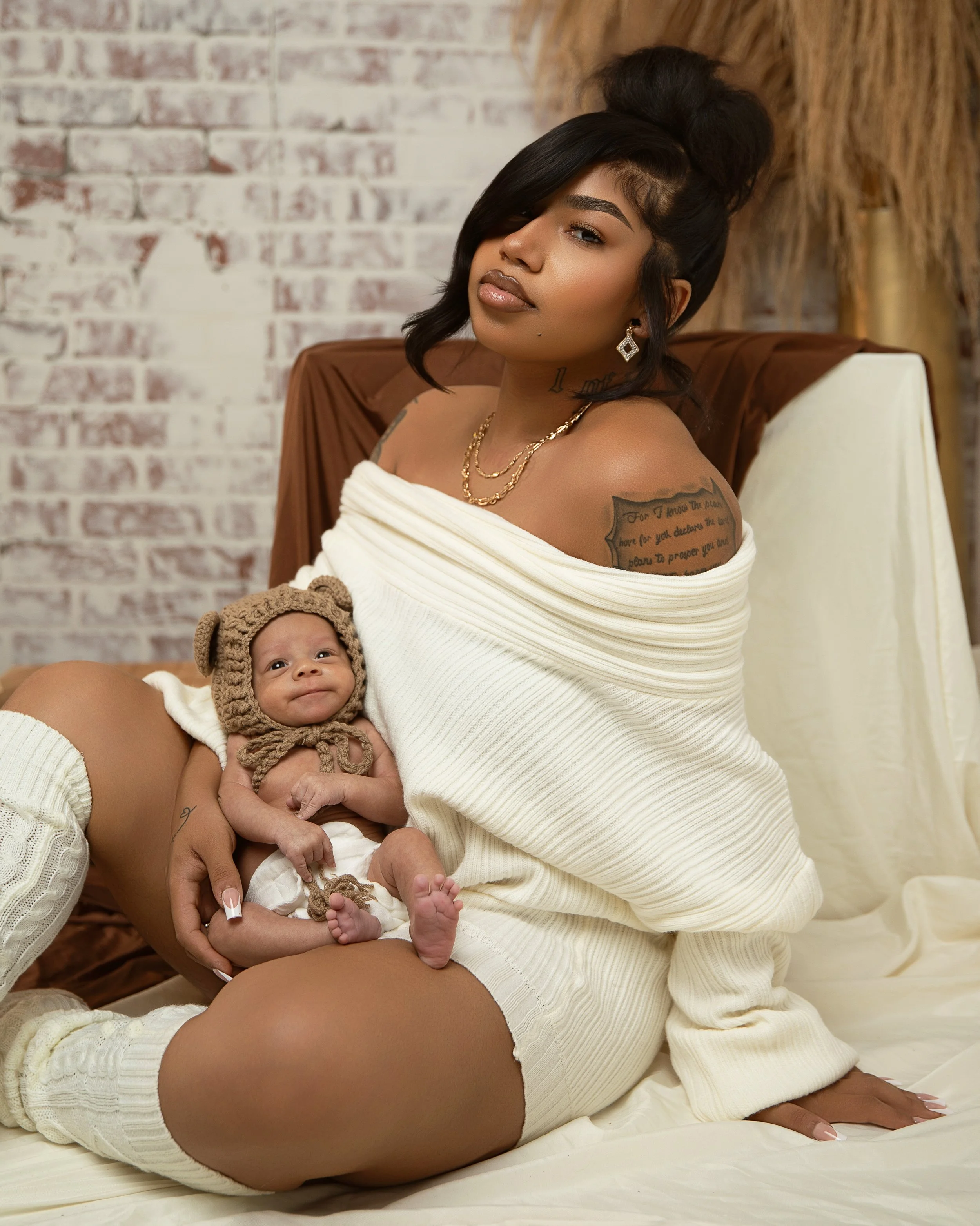 A woman with dark hair, wearing a white off-shoulder knitted sweater and thigh-high socks, sitting on a bed with a young baby dressed in a brown bear hat and white shorts, in a cozy bedroom with brick walls.