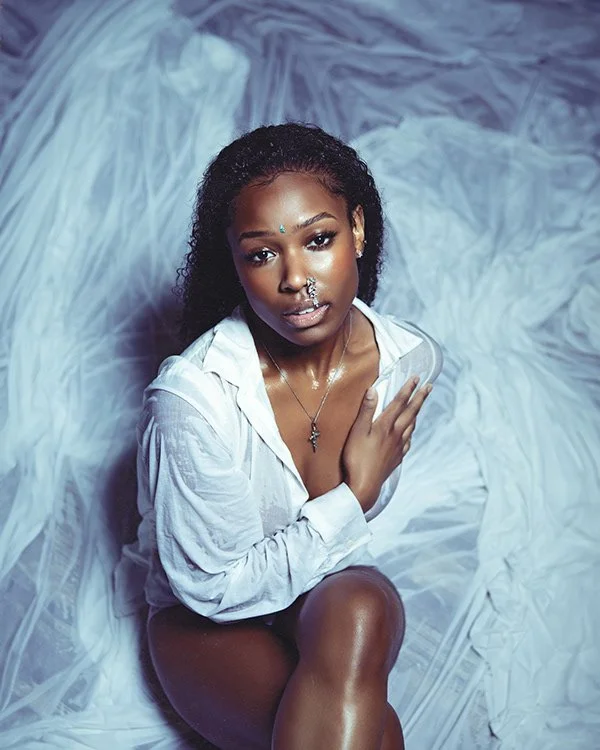 A woman with curly dark hair, wearing a white shirt, sitting on a white fabric background. She has jewelry on her face and neck and is looking at the camera.