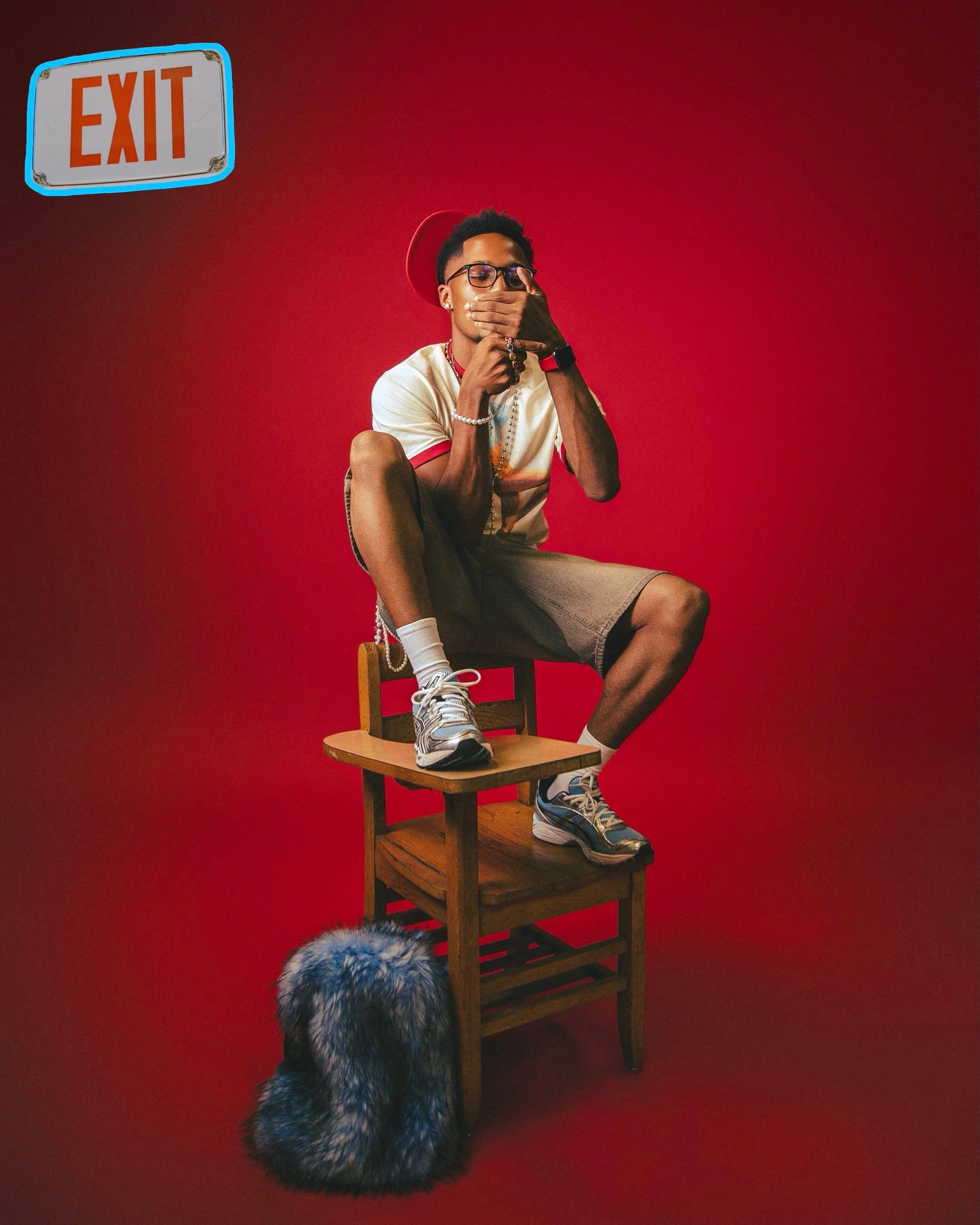 A young man wearing glasses, a white and red shirt, and brown shorts, sitting on a wooden chair with one foot on the seat and the other on the floor. He covers his mouth with his hand, wearing rings, a watch, and a bracelet. He is in front of a red background, with an illuminated exit sign in the upper left corner and a fuzzy blue and black fur object on the floor at the base of the chair.