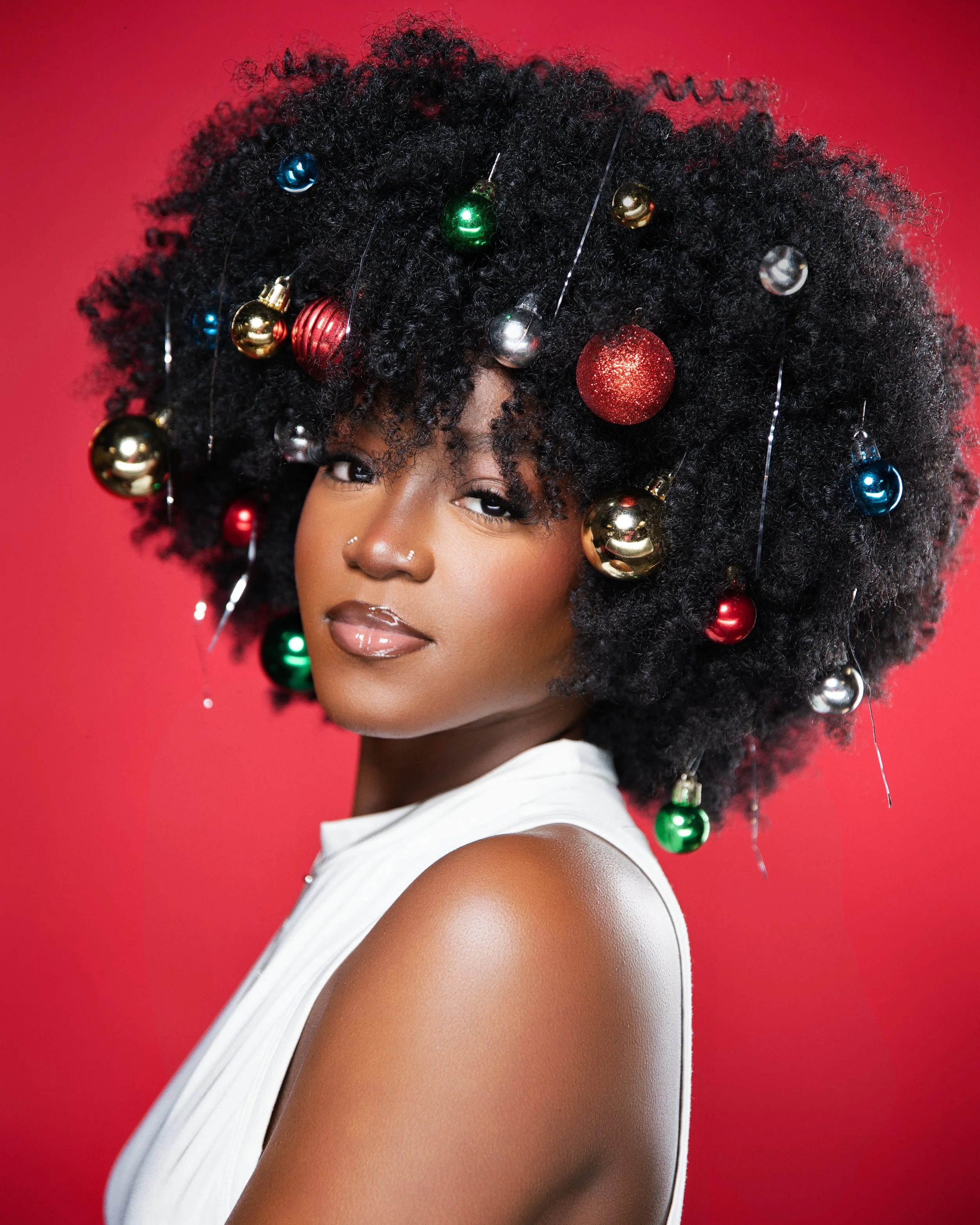 Woman with large, curly black hair decorated with colorful Christmas ornaments, wearing a white sleeveless top, against red background.
