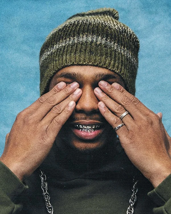 A man wearing a green knit beanie and a black shirt, covering his eyes with his hands, smiling with visible grills, and sporting rings on his fingers against a blue background.