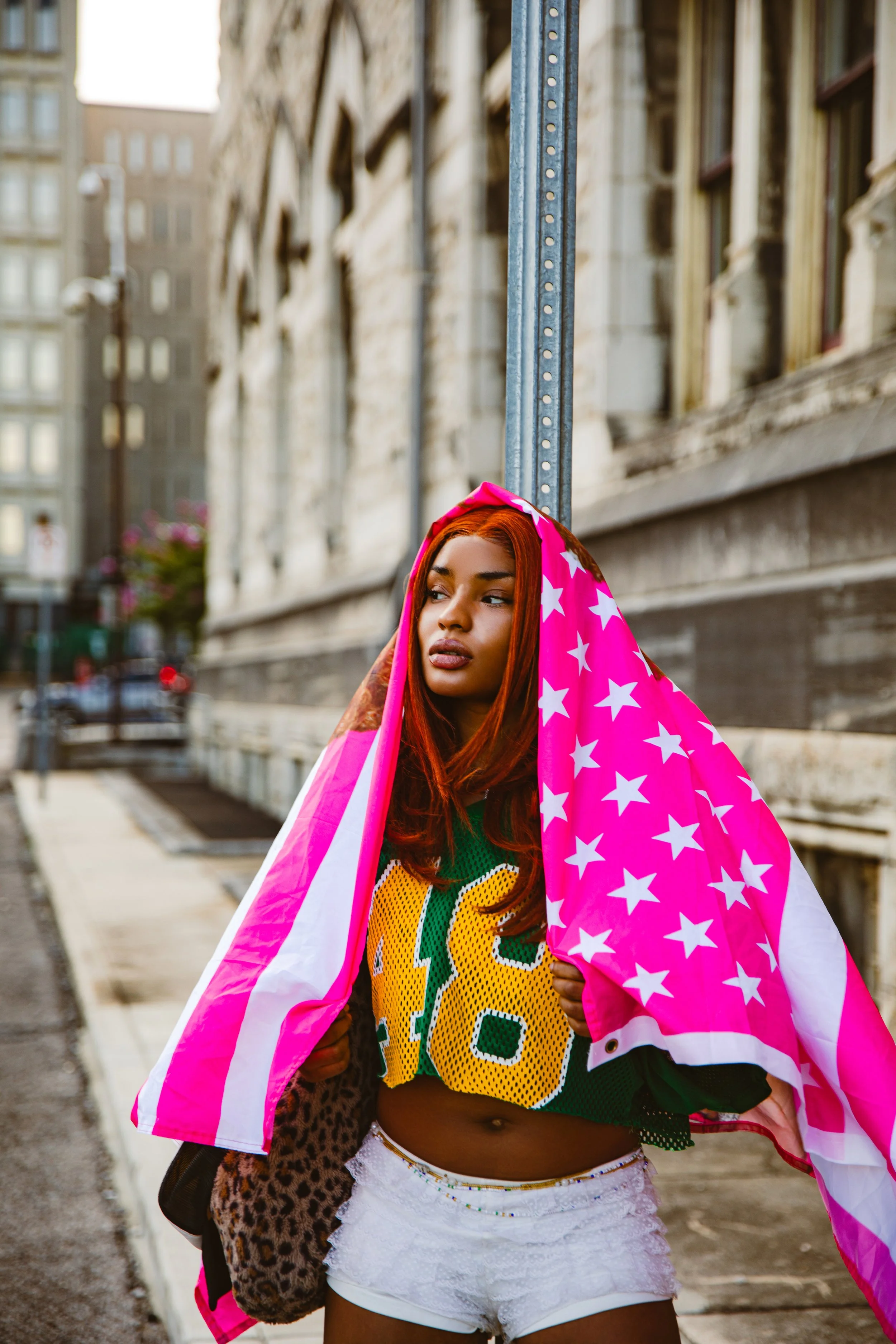A young woman with red hair standing on a city sidewalk, holding a pink star-patterned flag over her shoulders, wearing a green sports jersey with the number 18, white shorts, and a leopard-print bag.