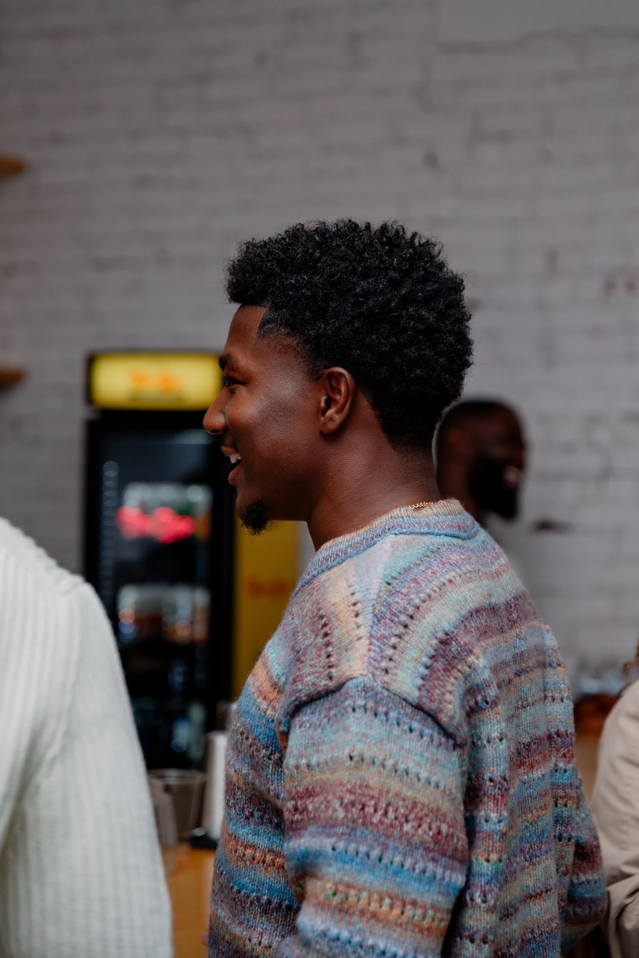 A smiling Black man with short curly hair wearing a multicolored striped sweater, standing in a casual indoor setting.