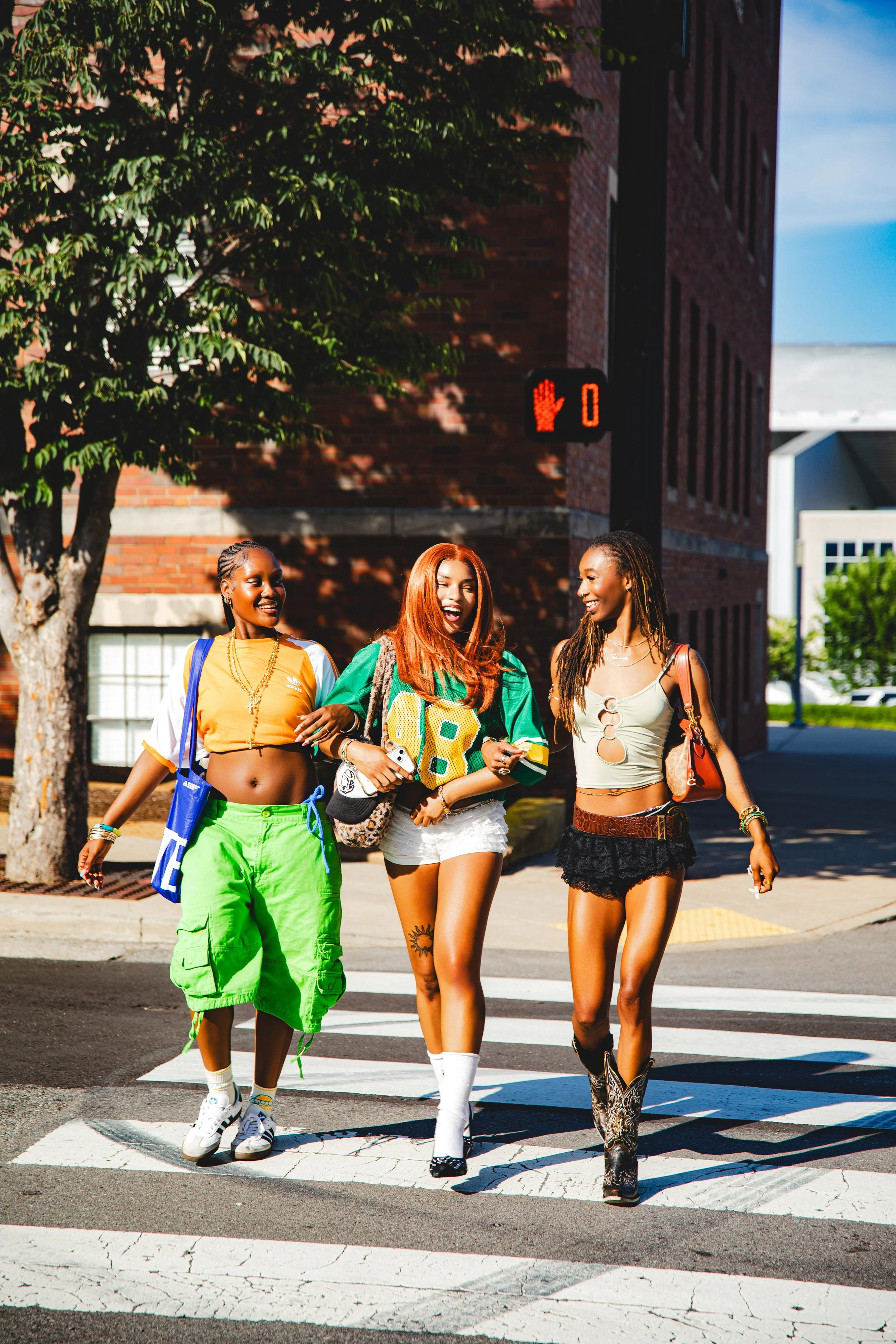 Three young women walking on crosswalk in urban area, smiling and having fun on sunny day, with red brick building and green trees in background.
